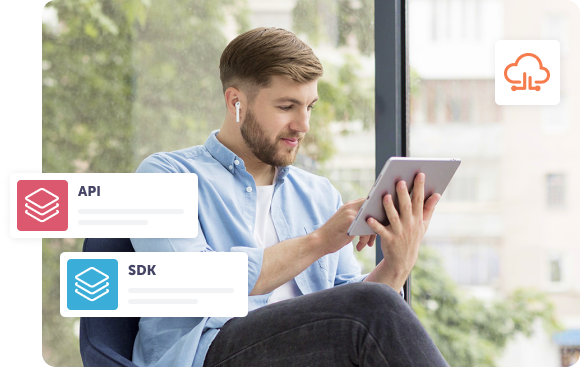 Vīrietis ar planšetdatoru sēž pie liela loga, ausīs ieliktas austiņas. Pārklājumā redzamas ikonas ar uzrakstiem “API” un “SDK”, kas izceļ programmatūras integrāciju, savukārt mākoņa ikona simbolizē mākoņdatošanas ieviešanu.