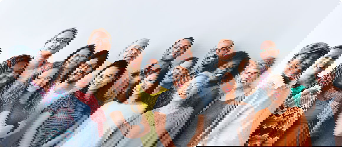 Attēlā redzama daudzveidīga, smaidīgu komandas locekļu grupa gaišā biroja telpā, kas simbolizē sadarbību, komandas darbu un pozitīvu darba vidi.