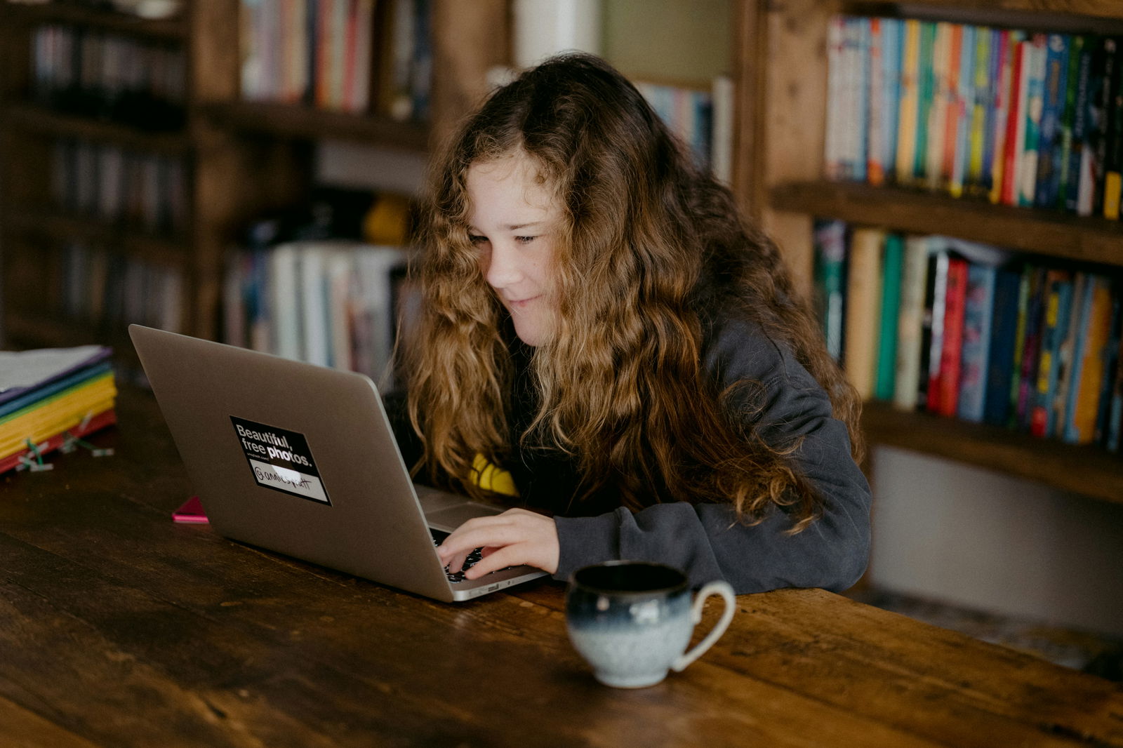 Girl with curly hair focused on laptop, coffee mug beside her, surrounded by books.