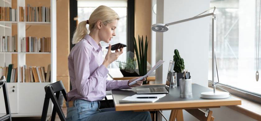 Una donna seduta a una scrivania con sopra un laptop utilizza uno smartphone.
