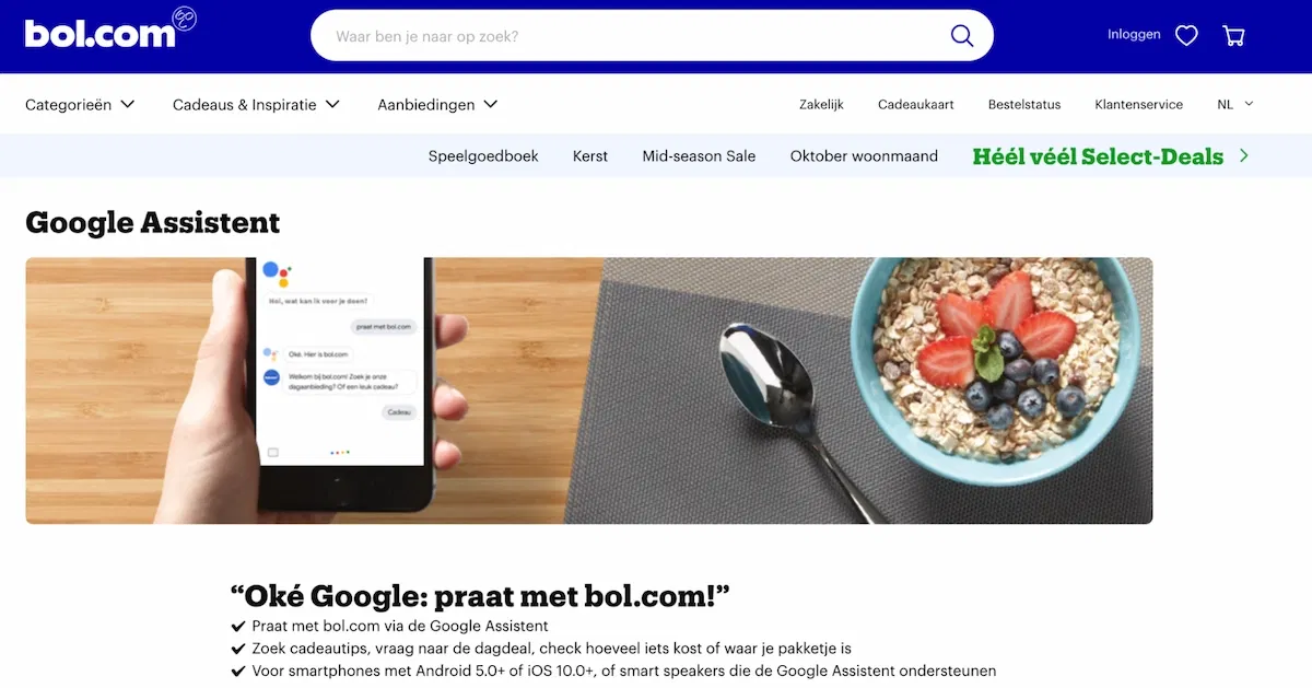 Screenshot der Website von bol.com mit der Seite zu Google Assistant. Das Bild zeigt eine Schale mit Müsli und eine Hand, die ein Smartphone hält, das über Google Assistant mit bol.com kommuniziert. Die Übersetzung des niederländischen Textes unter dem Bild lautet: Okay, Google: Sprich mit bol.com! Suchen Sie nach Geschenktipps, dem Deal des Tages, was etwas kostet oder wo Ihr Paket ist. Für Smartphones mit Android 5.0 oder höher, iOS 10.0 oder höher sowie für Smart Speaker, die Google Assistant unterstützen.