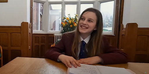 A young student in uniform smiles as she sits at a desk.