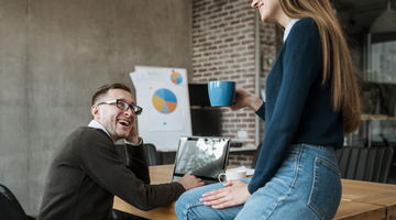Two colleagues having a friendly conversation in a modern office setting; a man is seated at a table with a laptop, smiling, while a woman sits on the table holding a blue coffee mug. A presentation with pie charts is visible in the background.