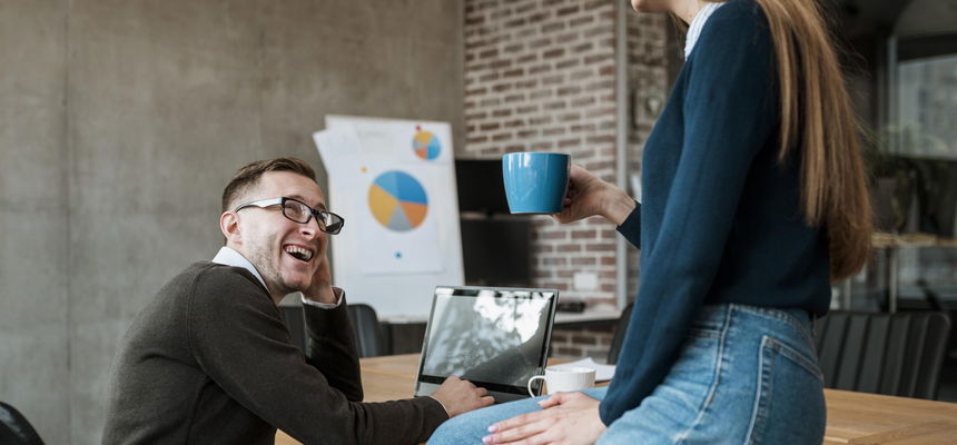 Two colleagues having a friendly conversation in a modern office setting; a man is seated at a table with a laptop, smiling, while a woman sits on the table holding a blue coffee mug. A presentation with pie charts is visible in the background.