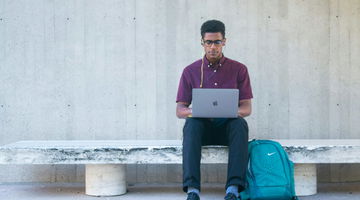 A man sitting on a bench with a laptop and blue backpack, wearing glasses and a purple shirt.
