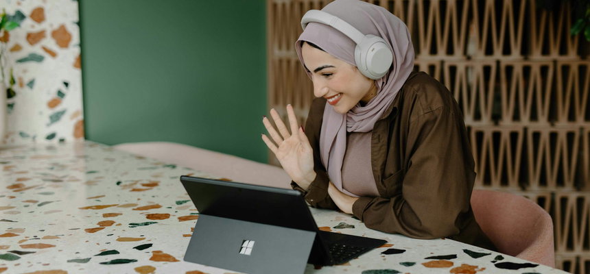 A woman wearing white headphones and a hijab, sitting at a table and waving her hand while watching a laptop.