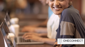 A woman smiles at the camera while typing on a laptop in a cafe. Warm ambient lighting and blurred background suggest a relaxed atmosphere. The text "ONECONNECT" is in the corner.