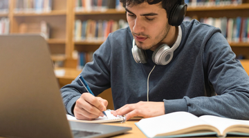 Student sitting a desk in a library, making notes, while wearing headphones, and with a laptop and books in front of him.