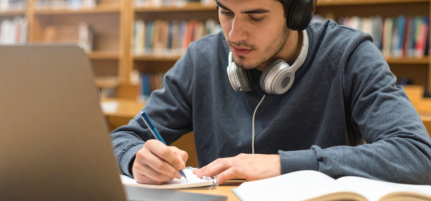 Student sitting a desk in a library, making notes, while wearing headphones, and with a laptop and books in front of him.