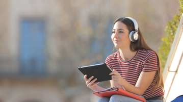 Girl with tablet wearing headphones sitting on a college campus building's stairs.