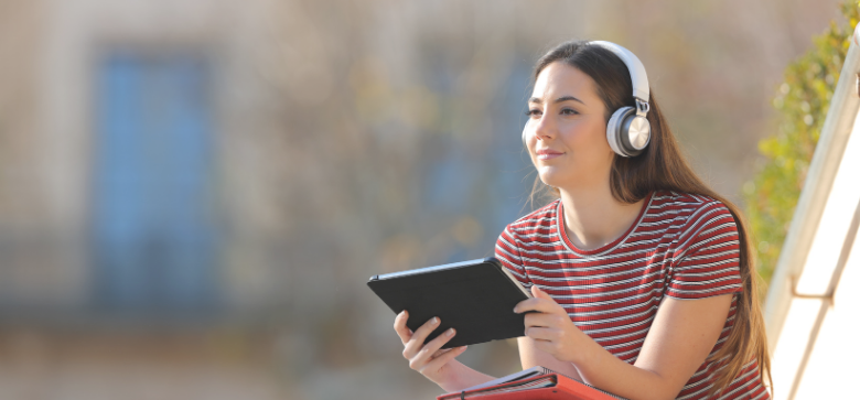 Girl with tablet wearing headphones sitting on a college campus building's stairs.
