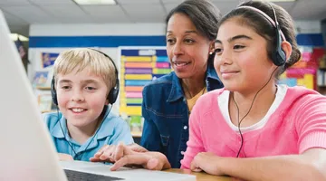 Dois alunos na escola usam fones de ouvido e tecnologia de conversão de texto em fala enquanto fazem seus trabalhos escolares em um computador portátil. O professor está sentado atrás deles, observando como trabalham.