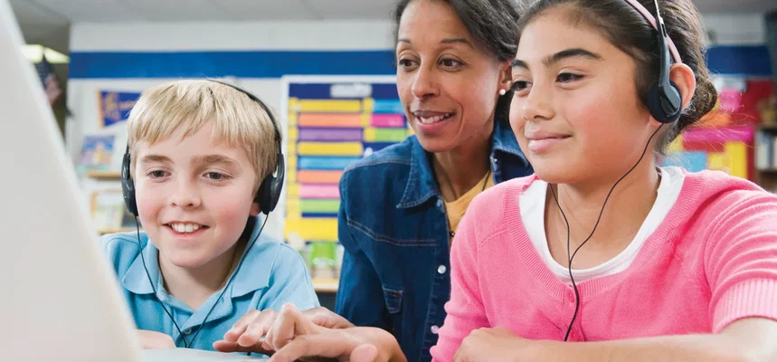 Dois alunos na escola usam fones de ouvido e tecnologia de conversão de texto em fala enquanto fazem seus trabalhos escolares em um computador portátil. O professor está sentado atrás deles, observando como trabalham.