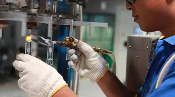 Un travailleur avec des gants et des lunettes de sécurité utilisant une torche à souder pour assembler des éléments en verre dans un atelier industriel.