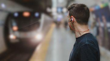 A wide horizontal banner featuring a man from the side, seen from the chest up, standing on a blurred subway platform as a train approaches in the background. He is wearing a dark t-shirt and bright orange wired earbuds. A subtle, white flowing wave sound line pattern emanates from his ear conveying a transport announcement, moving toward the left side of the frame across the subway station setting.