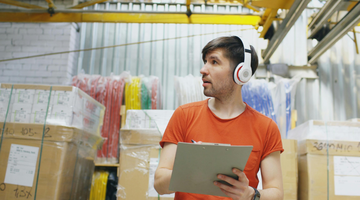 A young man in a clothing warehouse, with a rack of clothes behind him. He is dressed in an orange t-shirt, and is wearing headphones and holding a clipboard.