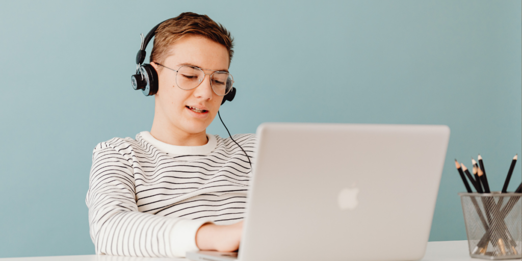 Image of a young man wearing a white and black striped t-shirt, sitting at a desk working at a laptop, and wearing headphones.