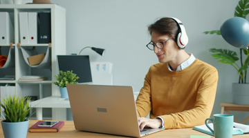 Young man with dark hair and glasses sitting in front of a laptop, and wearing headphones.