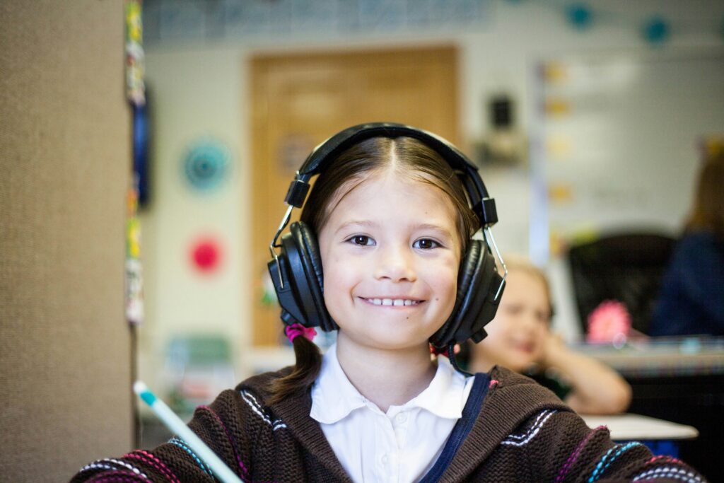 A young girl, around 7 or 8 years old, sitting in classroom. She is wearing headphones and smiling broadly, straight at the camera.