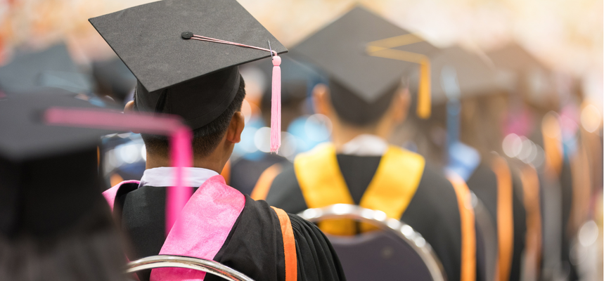 University students on graduation day, sitting in rows with their backs to the camera, wearing mortar boards and gowns.