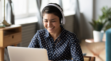 A smiling and smartly-dressed woman wearing a navy and white spotted blouse, sitting at a desk with a laptop and wearing headphones.