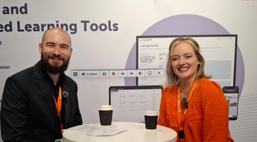 An image of two people, a man and a woman, sitting at a white pedestal table in front of a backdrop. The backdrop features text that includes "Learning Tools" and the name "Cirrus". The man is on the left, wearing a black blazer and an orange lanyard. The woman is on the right, wearing an orange top and a matching orange lanyard. Two coffee cups and some papers are on the table between them. In the background, there is a large screen displaying a digital interface with the text "Learning For Me" and a ReadSpeaker "Listen" button.