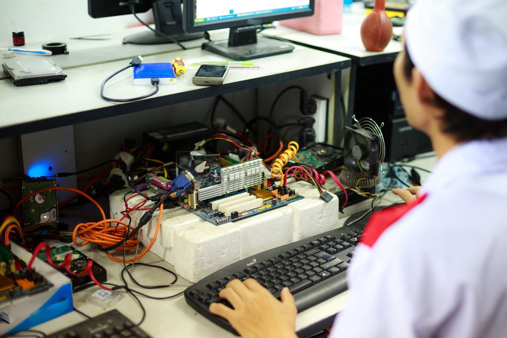 A technician working on a computer motherboard surrounded by wires and tools on a cluttered desk.