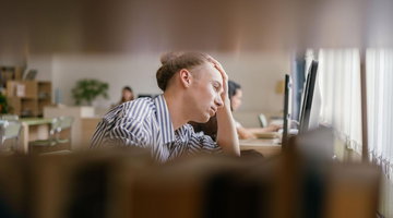 A man seated at a desk, focused on his computer screen, with a notepad and pen nearby. He looks focused, and his surroundings are blurry.