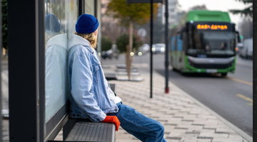 A person sits on a bench at a modern bus stop, looking toward a green bus approaching. Above the sidewalk, a digital passenger information display board shows real-time transit information.