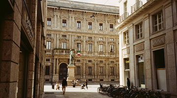 Una via stretta di Milano che si apre su una piccola piazza, dove una statua di bronzo su un basamento di pietra si trova davanti a un palazzo signorile con la bandiera italiana. Alcuni passanti attraversano la pavimentazione e una lunga fila di biciclette e parcheggiata sul lato destro.