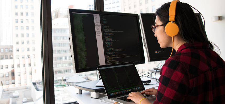 A developer wearing bright orange headphones works at a standing desk beside a city window, typing on a laptop in front of two large monitors filled with source code.