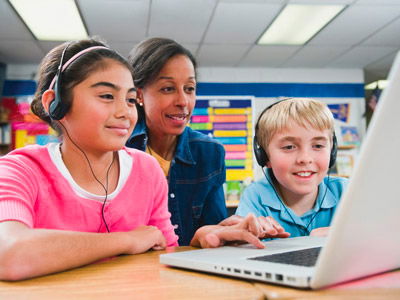 Two schoolchildren wearing headphones using text to speech as they do schoolwork on a laptop. The teacher is sitting behind them to see how they are doing.